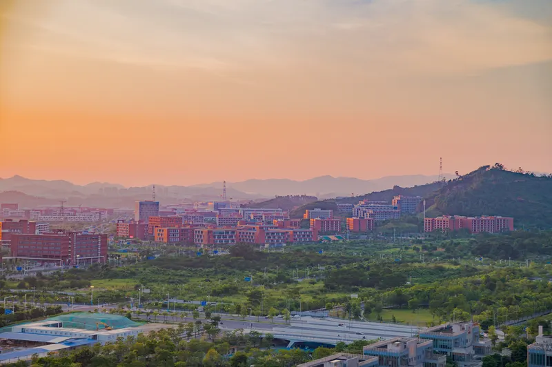 日落时分的城市远景，暖橙色的天空渐变到淡紫，红色的建筑群与绿化带在下方铺开